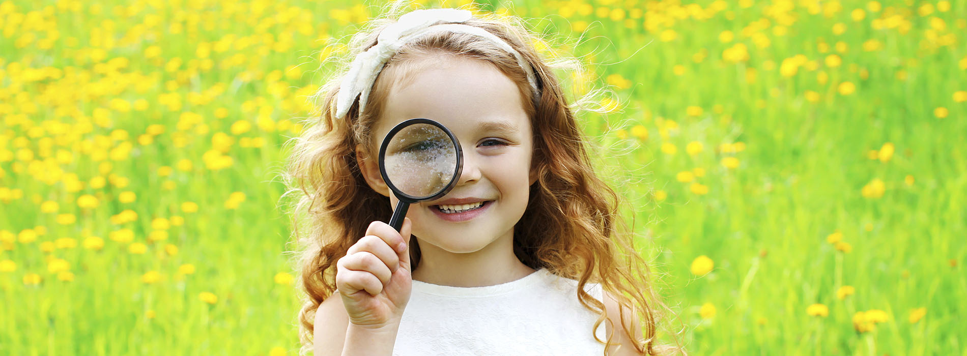 Smiling girl standing in a green field, looking through a magnifying glass